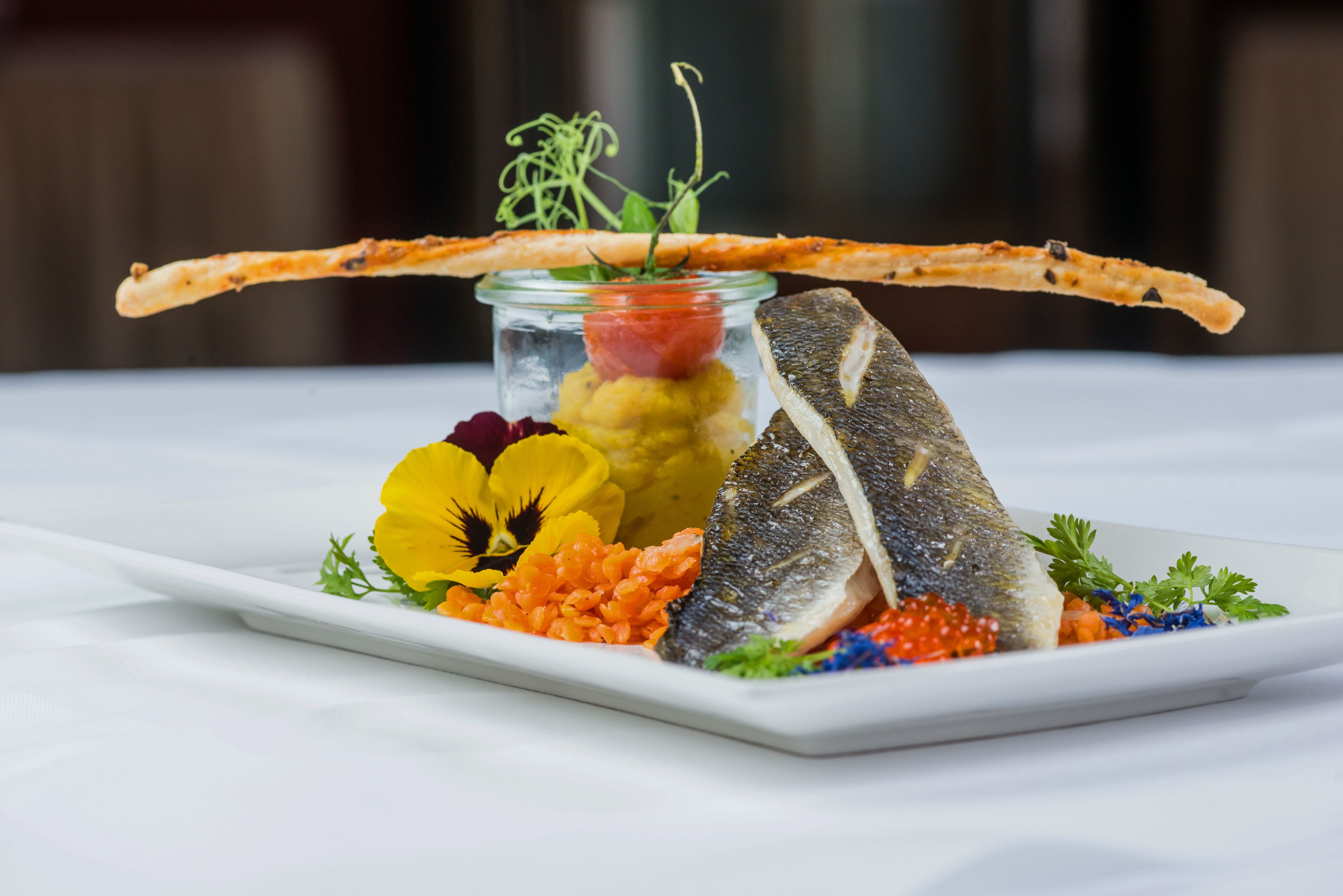 A white rectangular plate with a fish, caviar, and colorful vegetables on a white tablecloth.