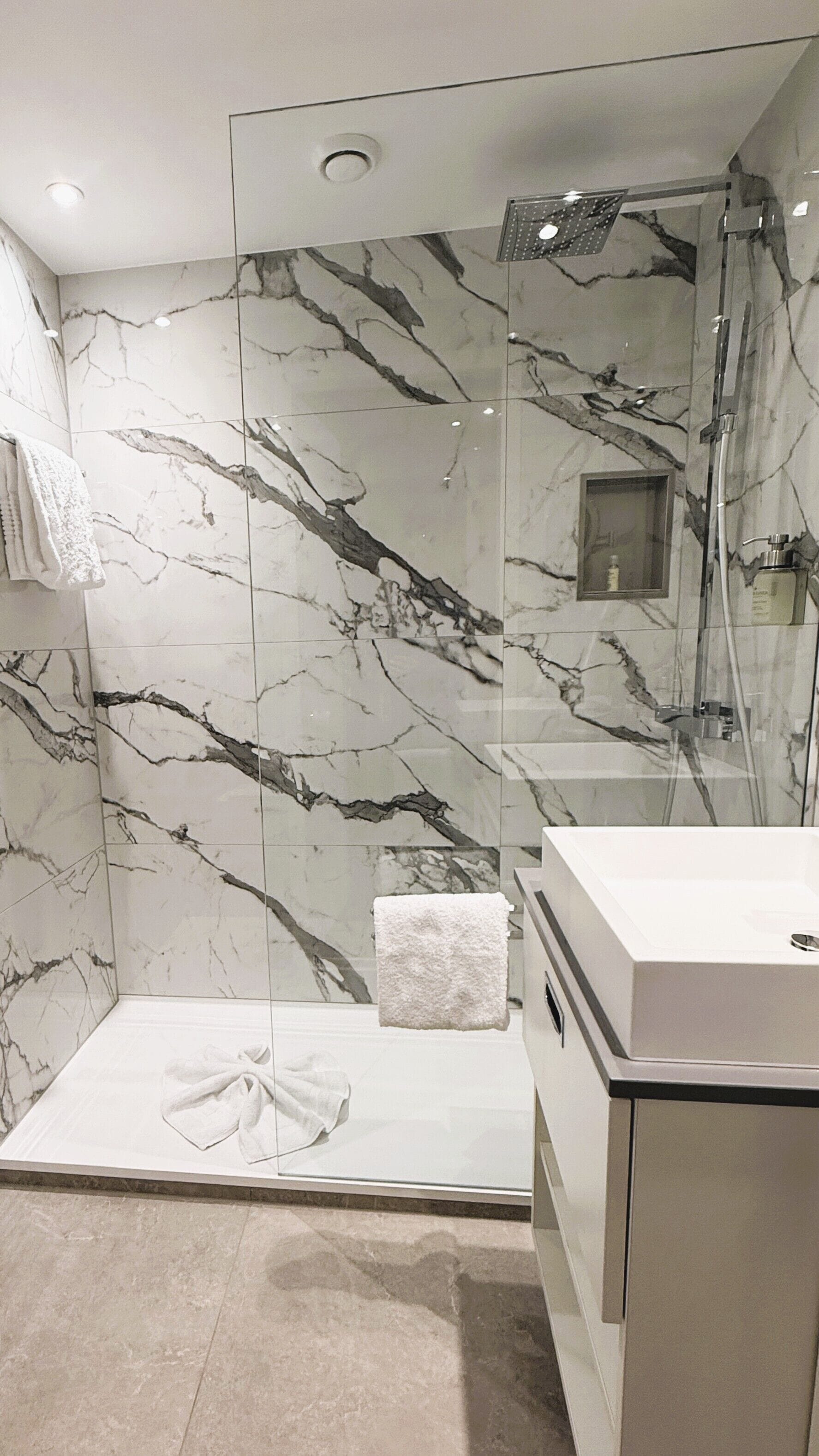 Un baño moderno con paredes de mármol, cabina de ducha de vidrio y lavamanos blanco.
