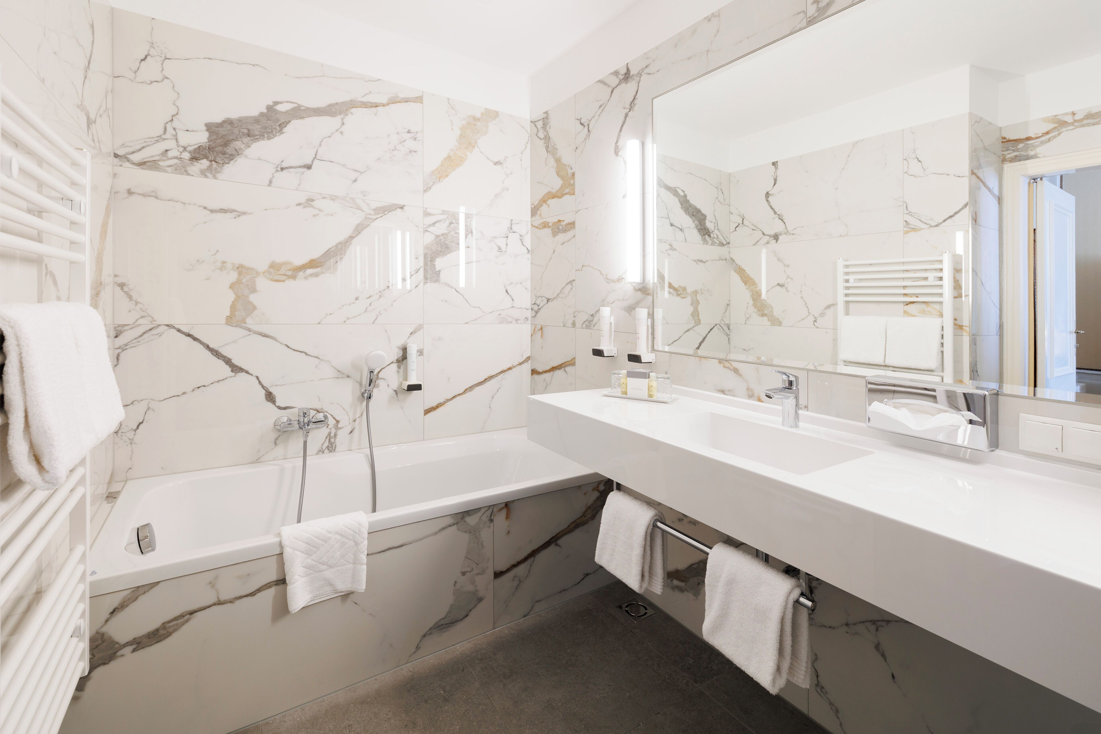 A modern bathroom with a marble wall, a bathtub, a large mirror, and a white countertop.