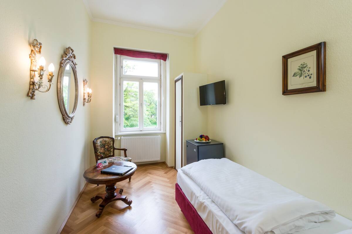 Een slaapkamer met een bed, bureau met laptop, stoel, raam met uitzicht, televisie en een lijstje aan de muur.