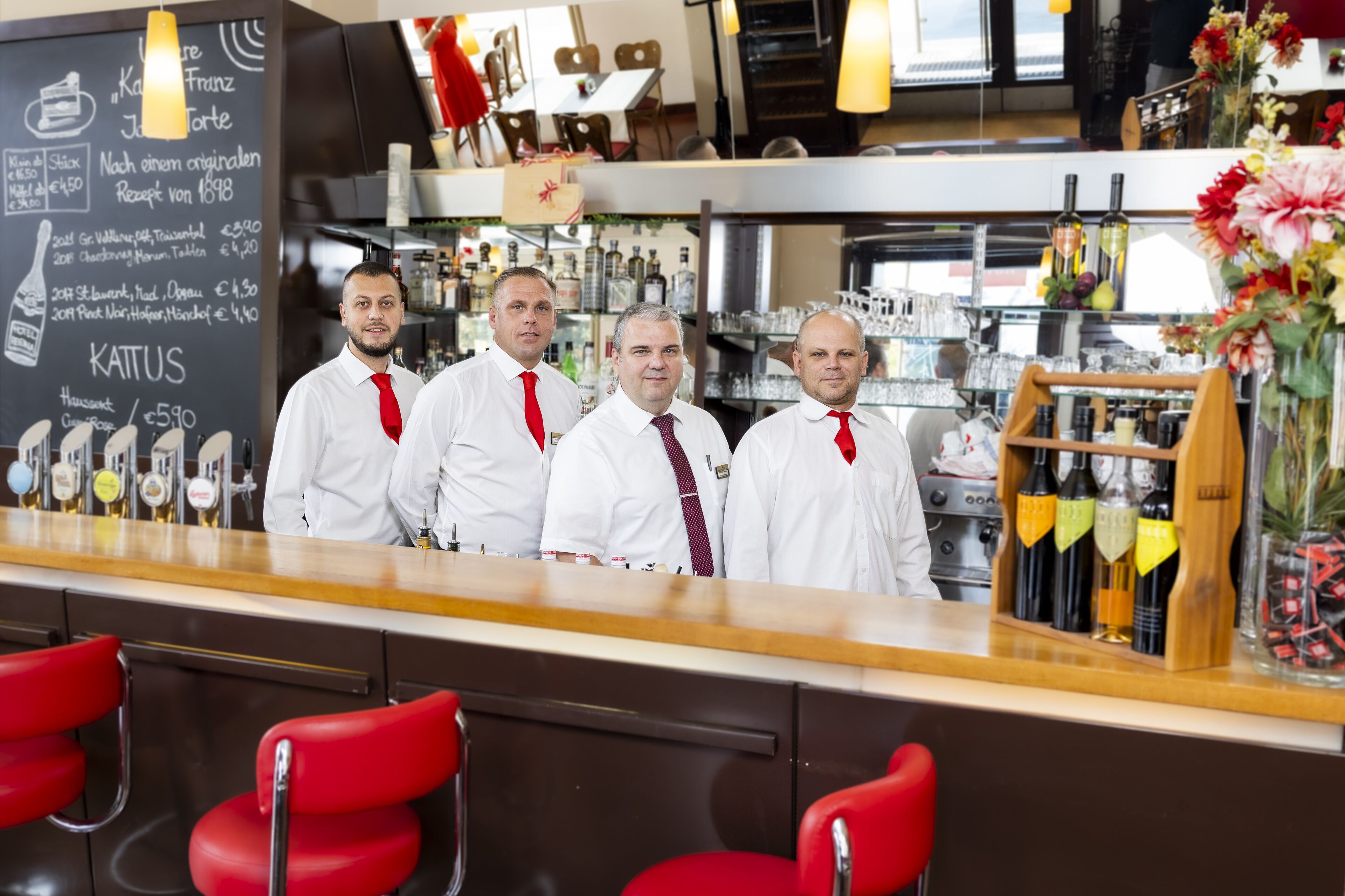 A bar area with four staff members standing behind the counter, a selection of bottles, and a chalkboard menu.