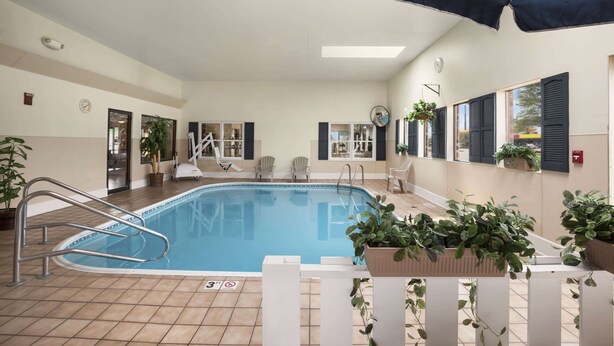 An indoor swimming pool with a tiled floor, a pool ladder, and a ceiling fan. There are potted plants and a white fence around the pool. - Days Inn by Wyndham Oglesby/ Starved Rock (Oglesby)