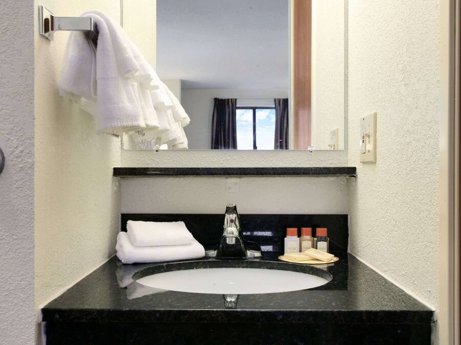 Baño de hotel con lavamanos de superficie negra, espejo encima y toallero con una toalla doblada.