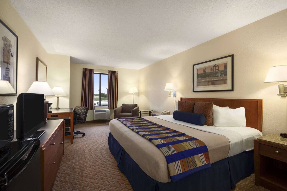 Una habitación de hotel con una cama grande, un escritorio con televisor, una silla, una lámpara de escritorio y una ventana con cortinas.