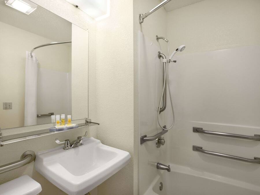 Ein Badezimmer mit Waschbecken, Spiegel und Dusche. An der Duschtür befinden sich Haltegriffe und oberhalb des Waschbeckens ein Handtuchhalter.