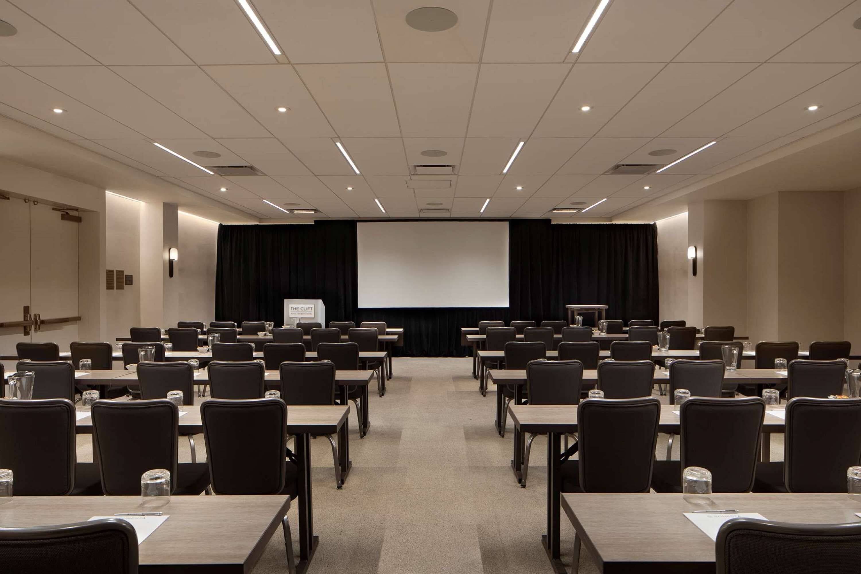 Una sala conferenze con file di tavoli e sedie, uno schermo per proiezioni e un palco con un podio.
