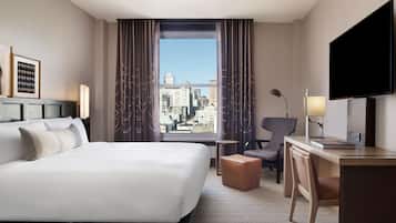 Habitación de hotel con una cama grande, un escritorio con silla, televisión y vista a la ciudad por la ventana.