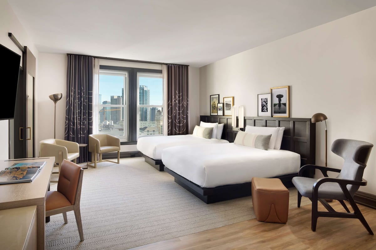Une chambre d’hôtel moderne avec un grand lit, deux tables de chevet, un bureau et une chaise. La fenêtre offre une vue sur la ville.