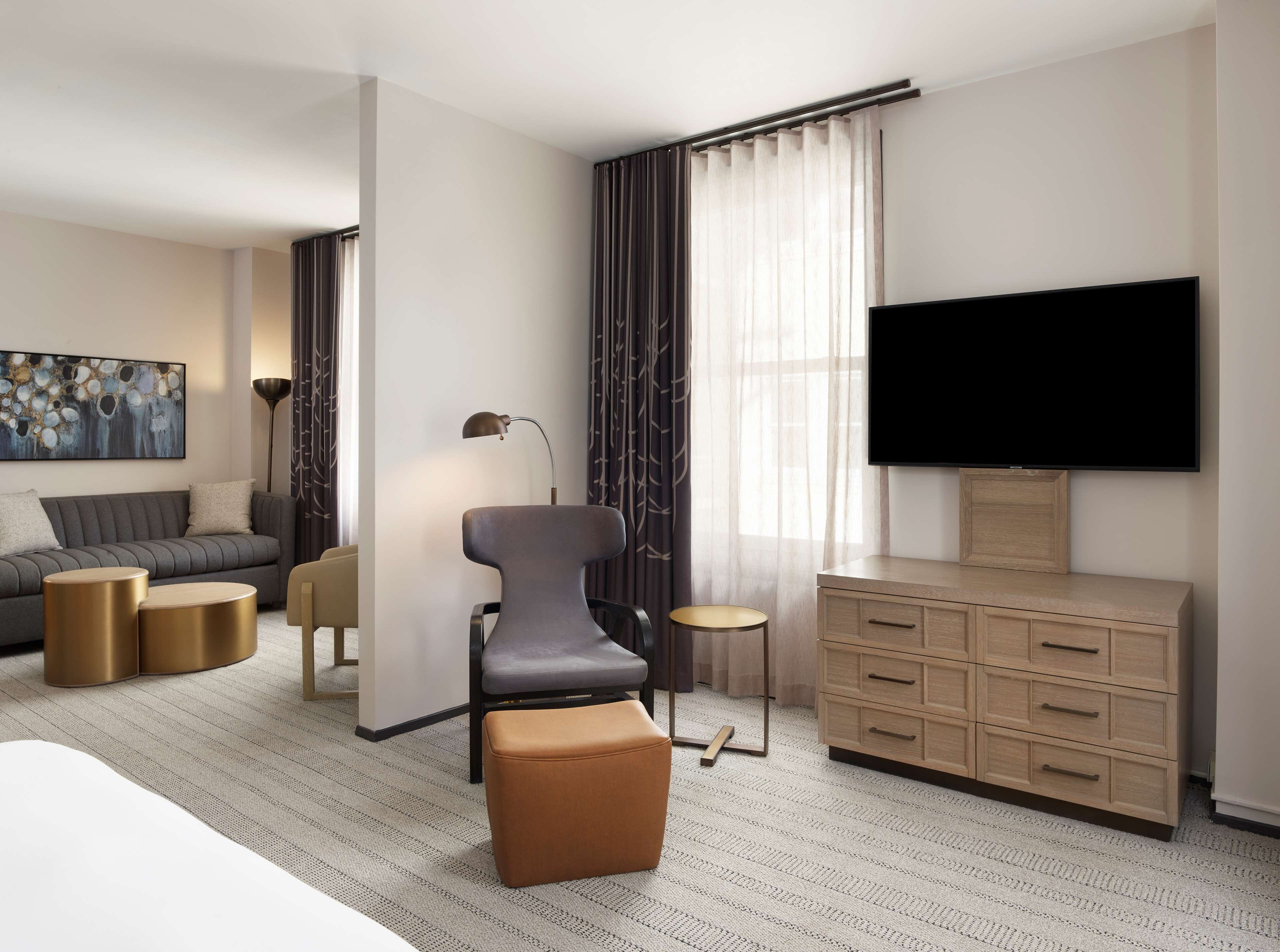 Una habitación de hotel moderna con sofá, sillón, reposapiés, televisor y cómoda de madera.