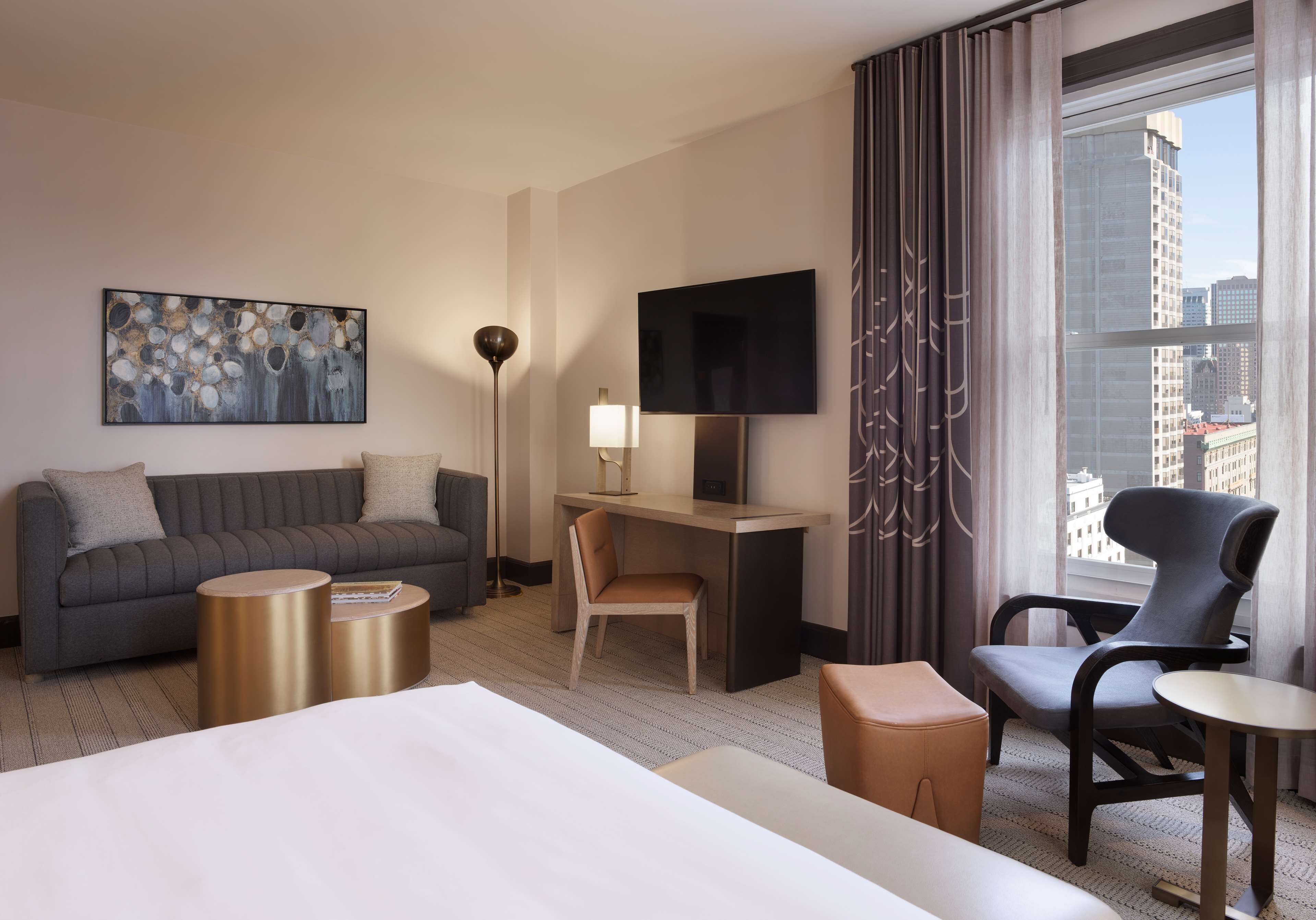 A hotel room with a bed, a sofa, a desk, a chair, a TV, and a large window with a city view.