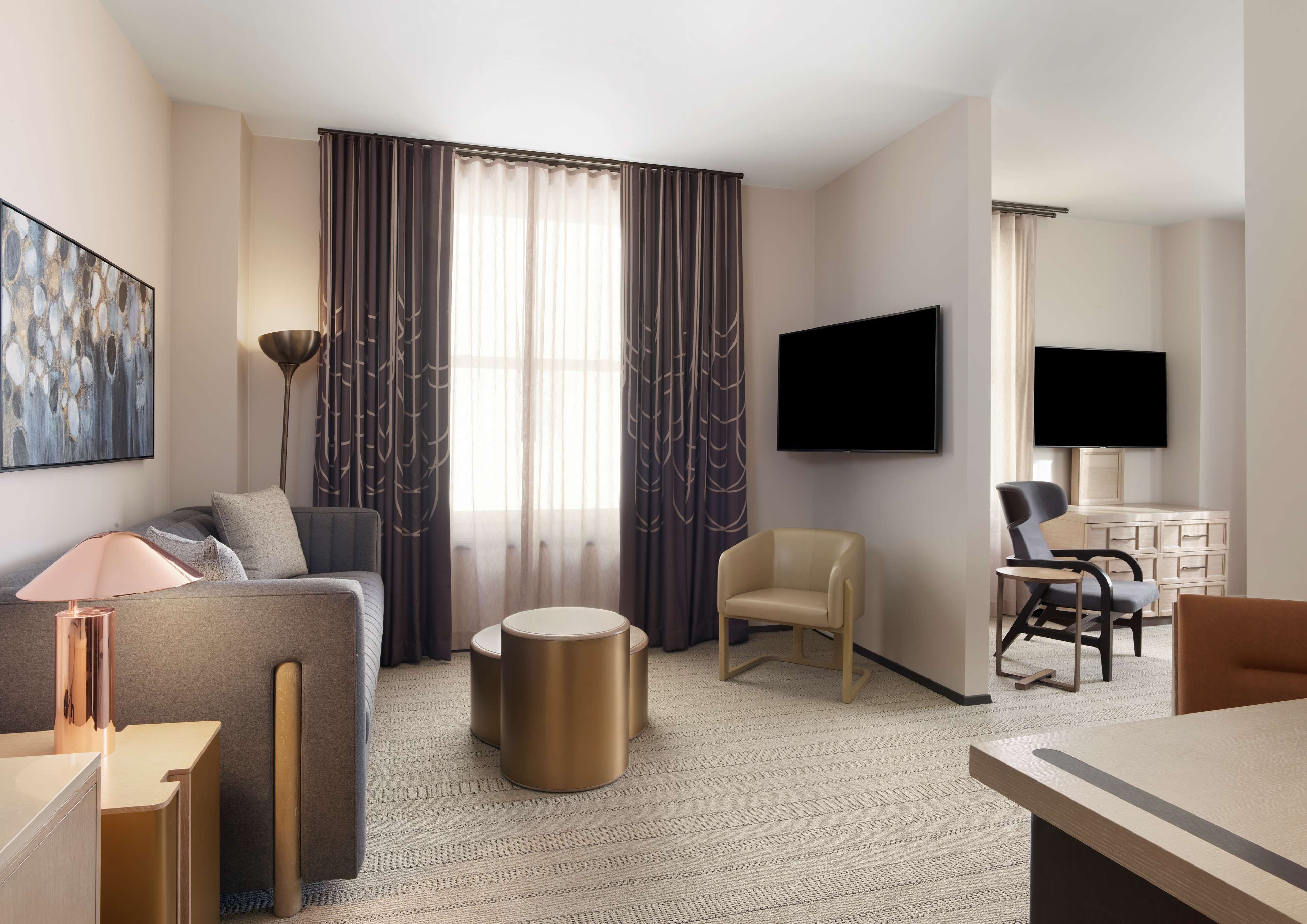 A modern hotel room with a sofa, two armchairs, a coffee table, a TV, and a desk.