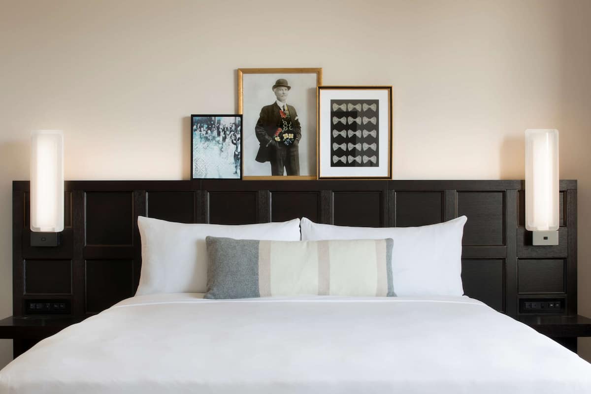 Une chambre d’hôtel avec un grand lit, deux tables de chevet, une tête de lit dotée de lampes fixées au mur et des tableaux encadrés accrochés au mur.