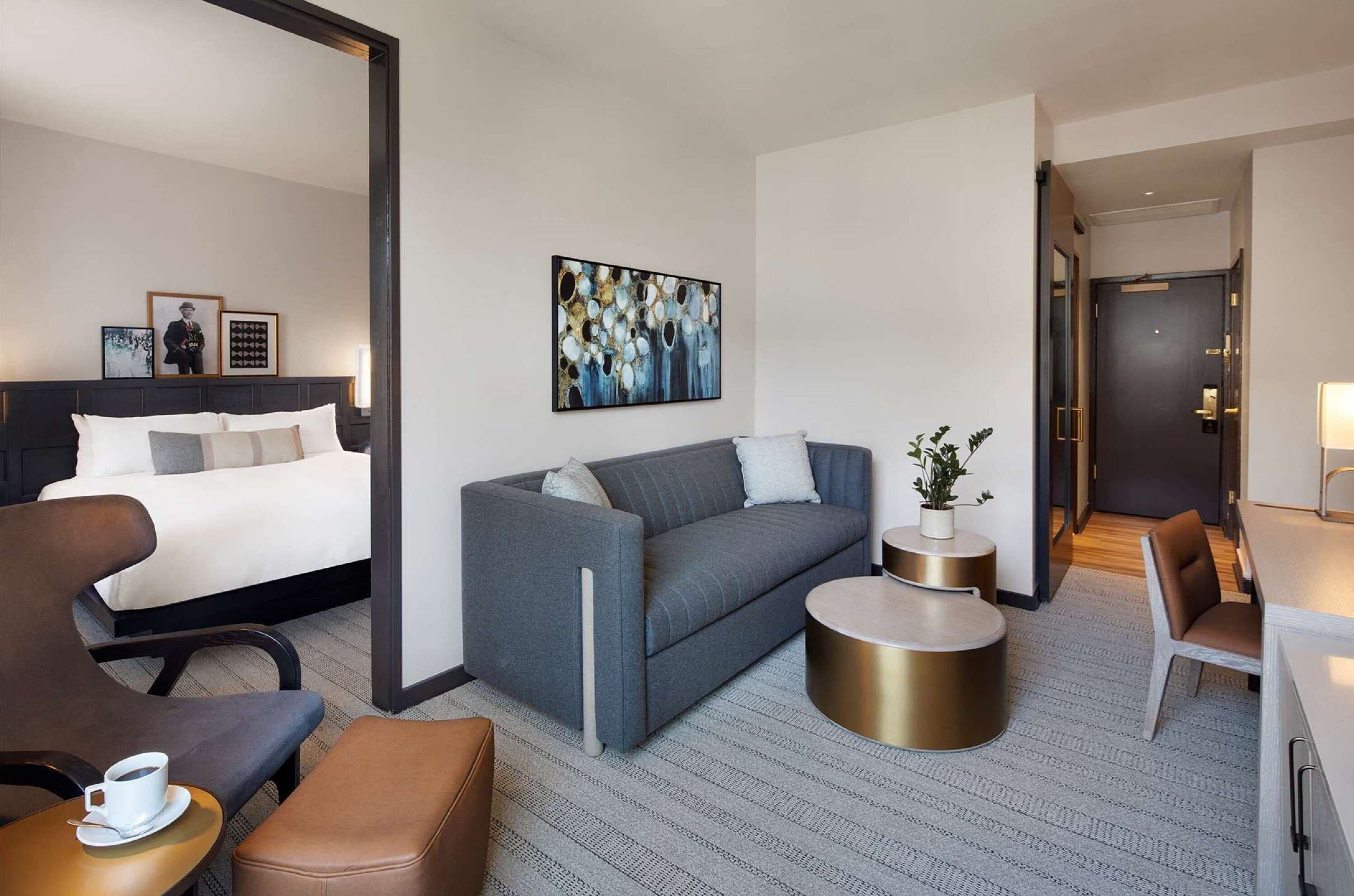 A modern hotel room with a bed, a grey sofa, a coffee table, and a desk with a chair.