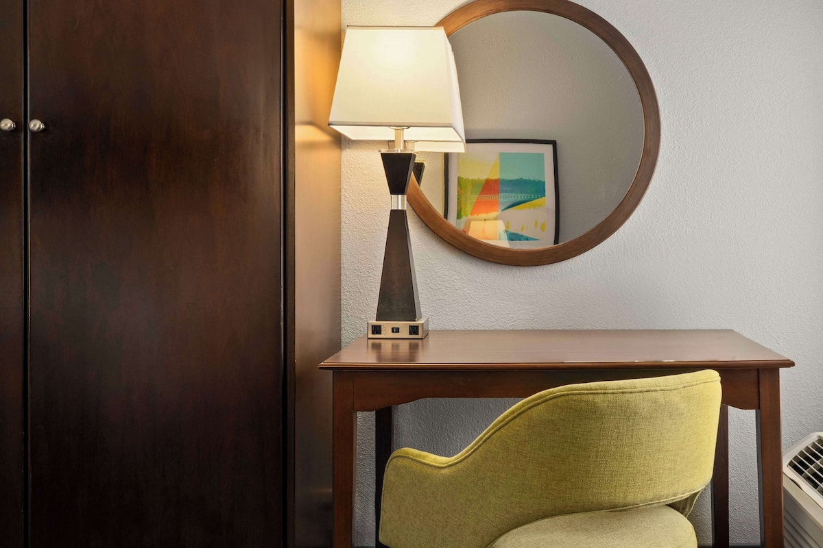 Una camera d'albergo con un armadio in legno, una scrivania con una lampada, una sedia e uno specchio tondo appeso al muro.