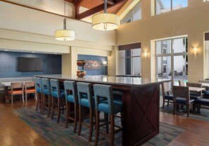 A spacious bar area with a long counter and multiple stools, a flat-screen TV mounted on the wall, and a large window providing a view of the outside. - Hampton Inn & Suites Newport News (Oyster Point) (Newport News)