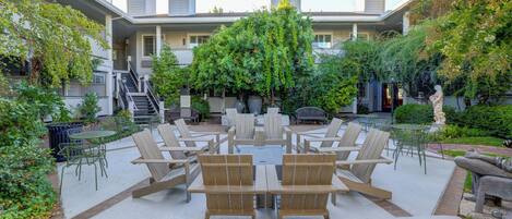 Un patio al aire libre con mesas y sillas de madera, rodeado de vegetación y un edificio al fondo.