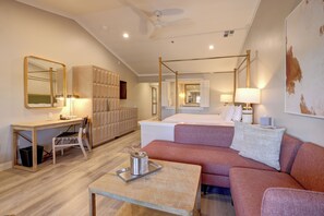 A modern hotel room with a four-poster bed, a sitting area with a sofa and a small table, a desk with a chair, and a large mirror.