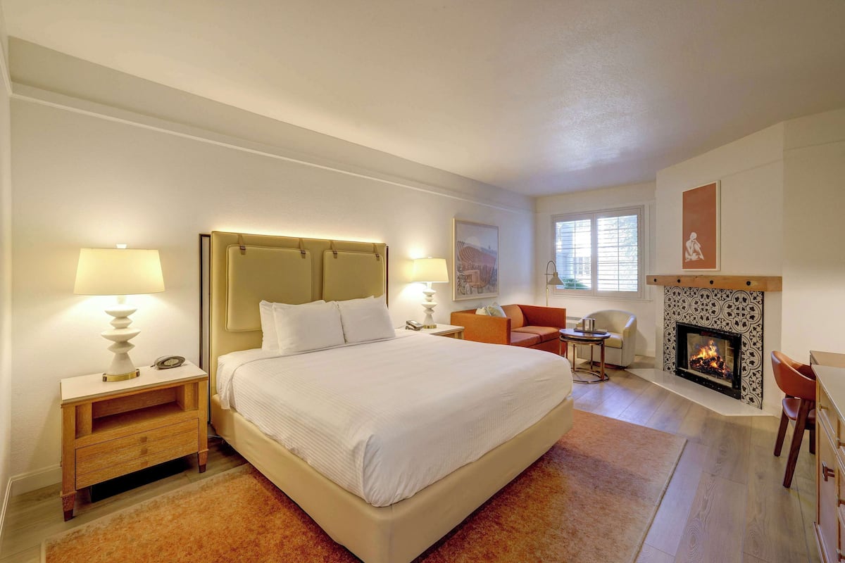 Habitación de hotel con una cama grande, lámparas de noche, chimenea y una zona de estar con una silla y una mesa.