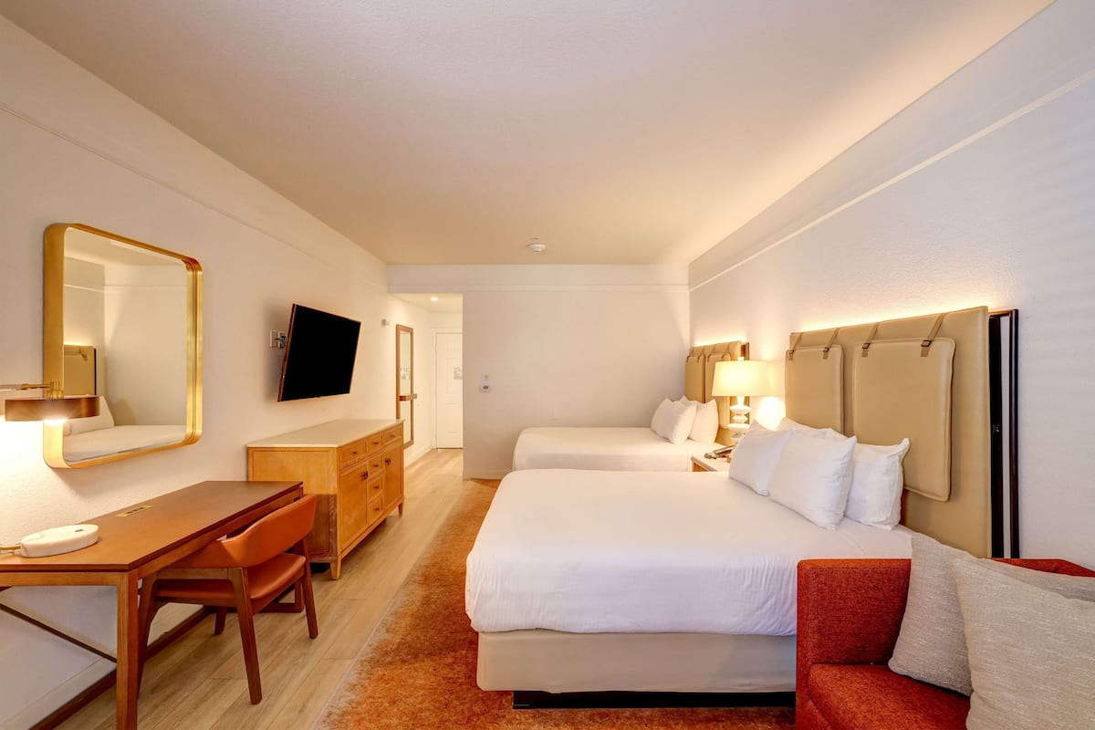 Una habitación de hotel con dos camas, un escritorio con silla, un televisor y un espejo.