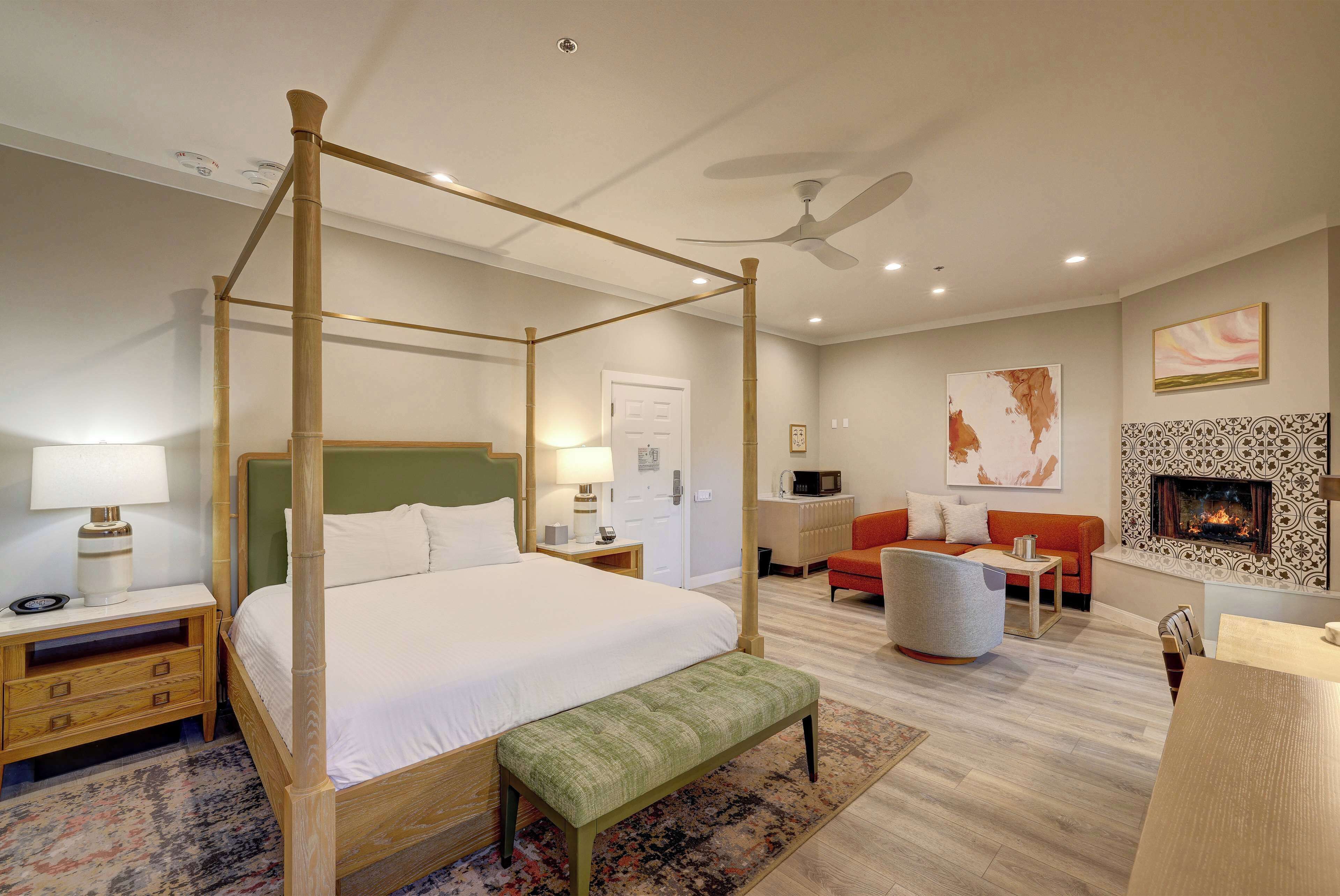 A four-poster bed with a bench, a sofa, a fireplace, and a ceiling fan in a well-lit room with a patterned rug.