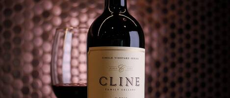 A bottle of Cline wine and a glass of red wine on a table with a textured background.