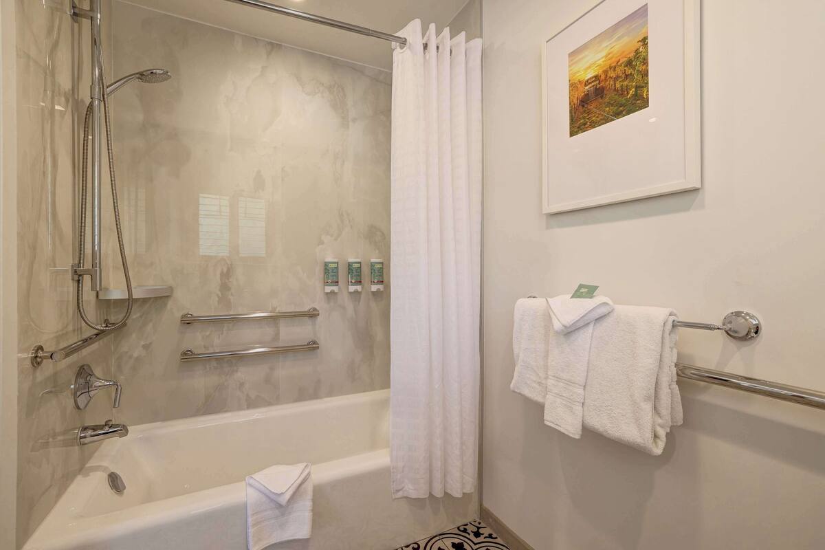 Un baño con bañera, ducha y barras de apoyo. Hay toallas en un perchero y un cuadro en la pared.