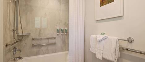 A bathroom with a bathtub, shower, and handrails. There are towels on a rack and a framed picture on the wall.