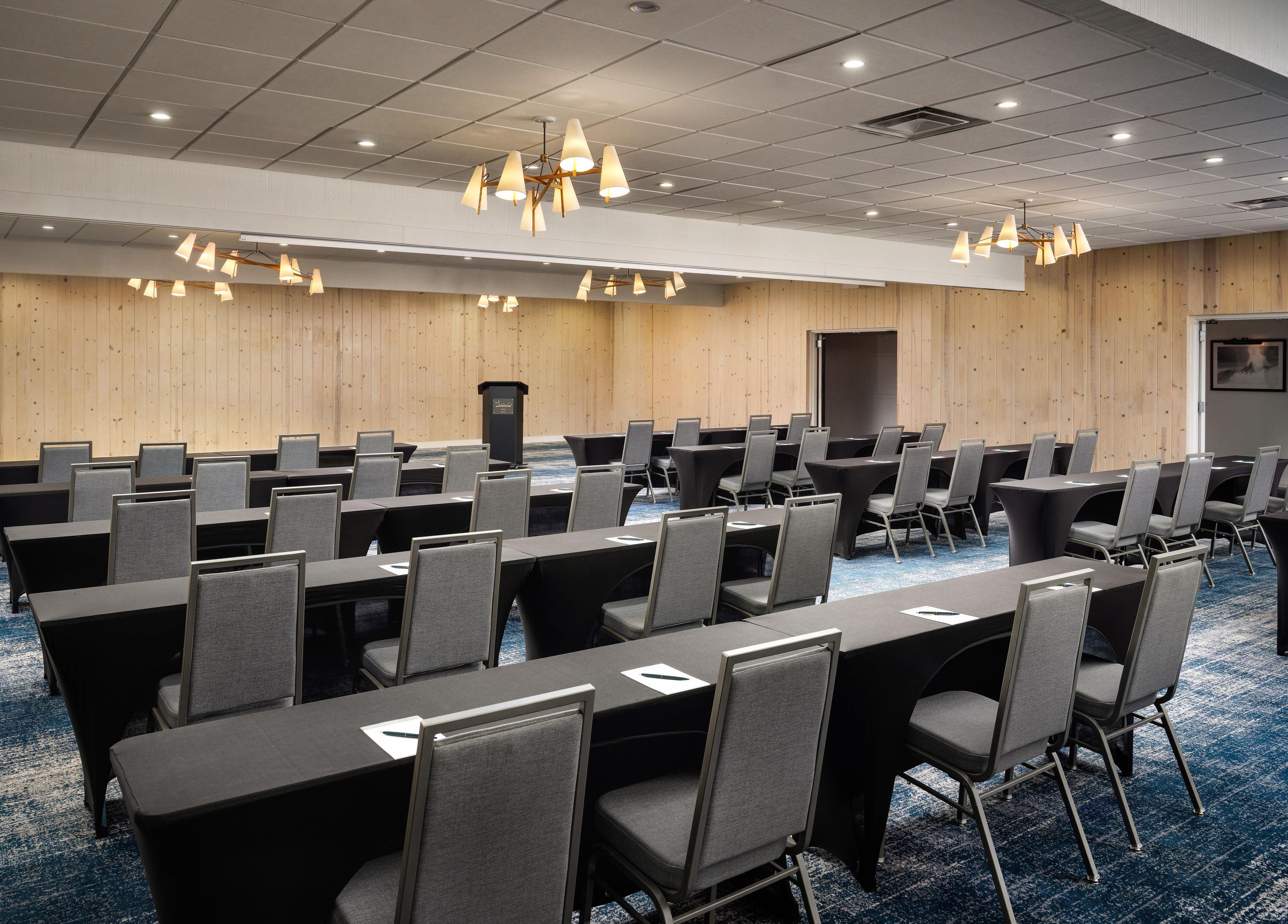 A conference room with rows of tables and chairs, a podium, and ceiling lights.