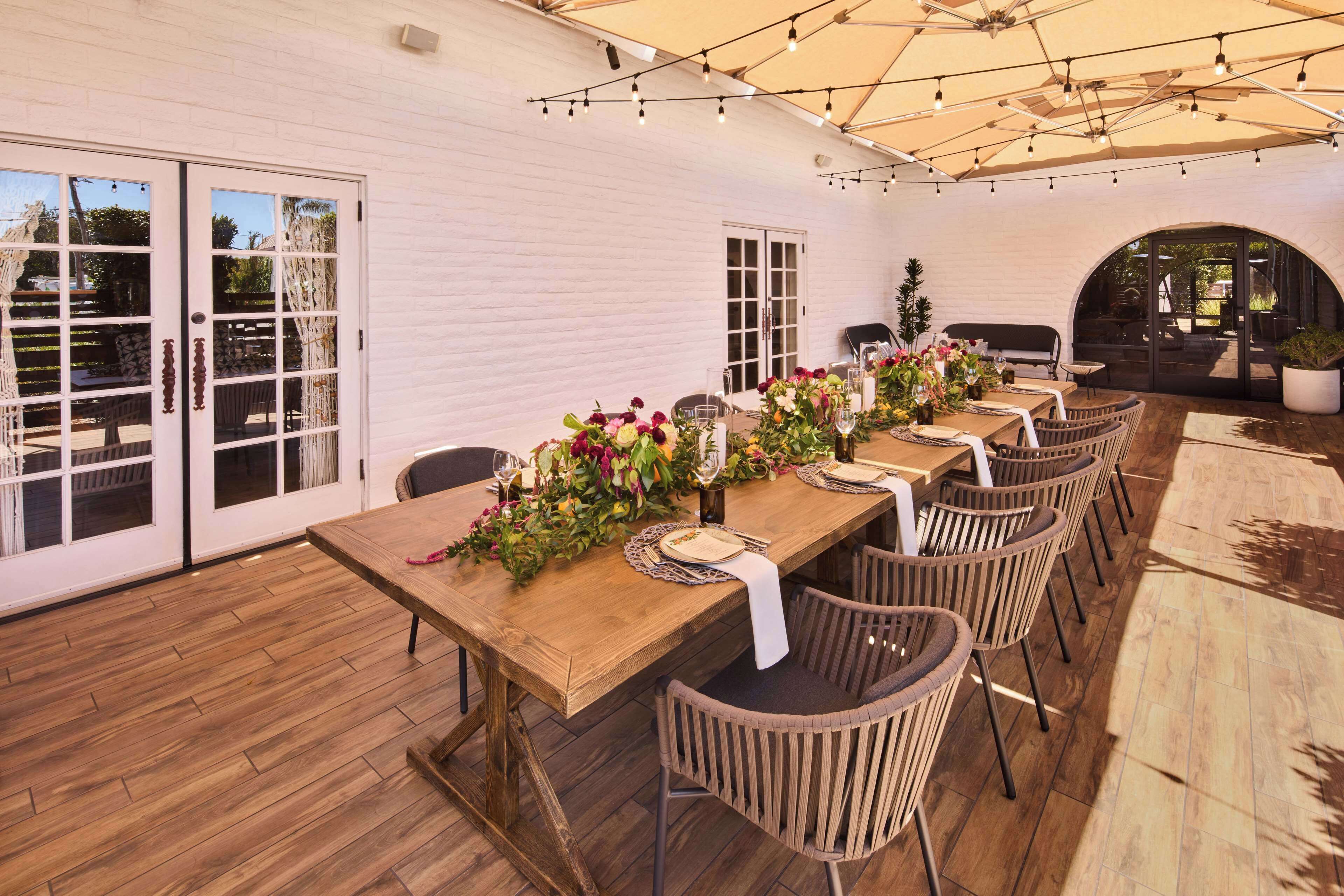 An outdoor dining area with wooden tables and chairs, a white brick wall, and string lights.