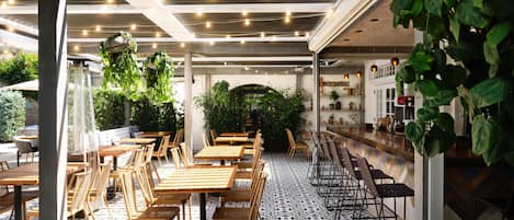 Un comedor interior con mesas y sillas de madera, piso estampado y plantas colgantes.