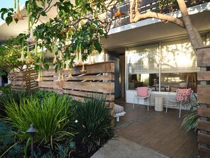 Un patio exterior moderno con macetas de madera, una zona de estar con sillas blancas y vistas del interior a través de amplios ventanales.