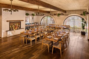 A spacious dining area with wooden tables and chairs, a brick wall, and arched windows with decorative curtains. - The Leta Santa Barbara Goleta, Tapestry Collection by Hilton (Goleta)