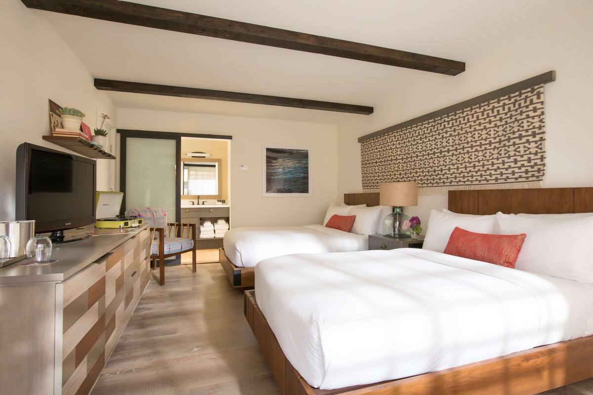 Une chambre d’hôtel avec deux lits, une télévision, un bureau avec un ordinateur et une salle de bain visible à travers une porte ouverte.