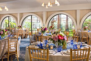 Un salón de banquetes con mesas redondas, sillas de madera y centros de mesa con flores.