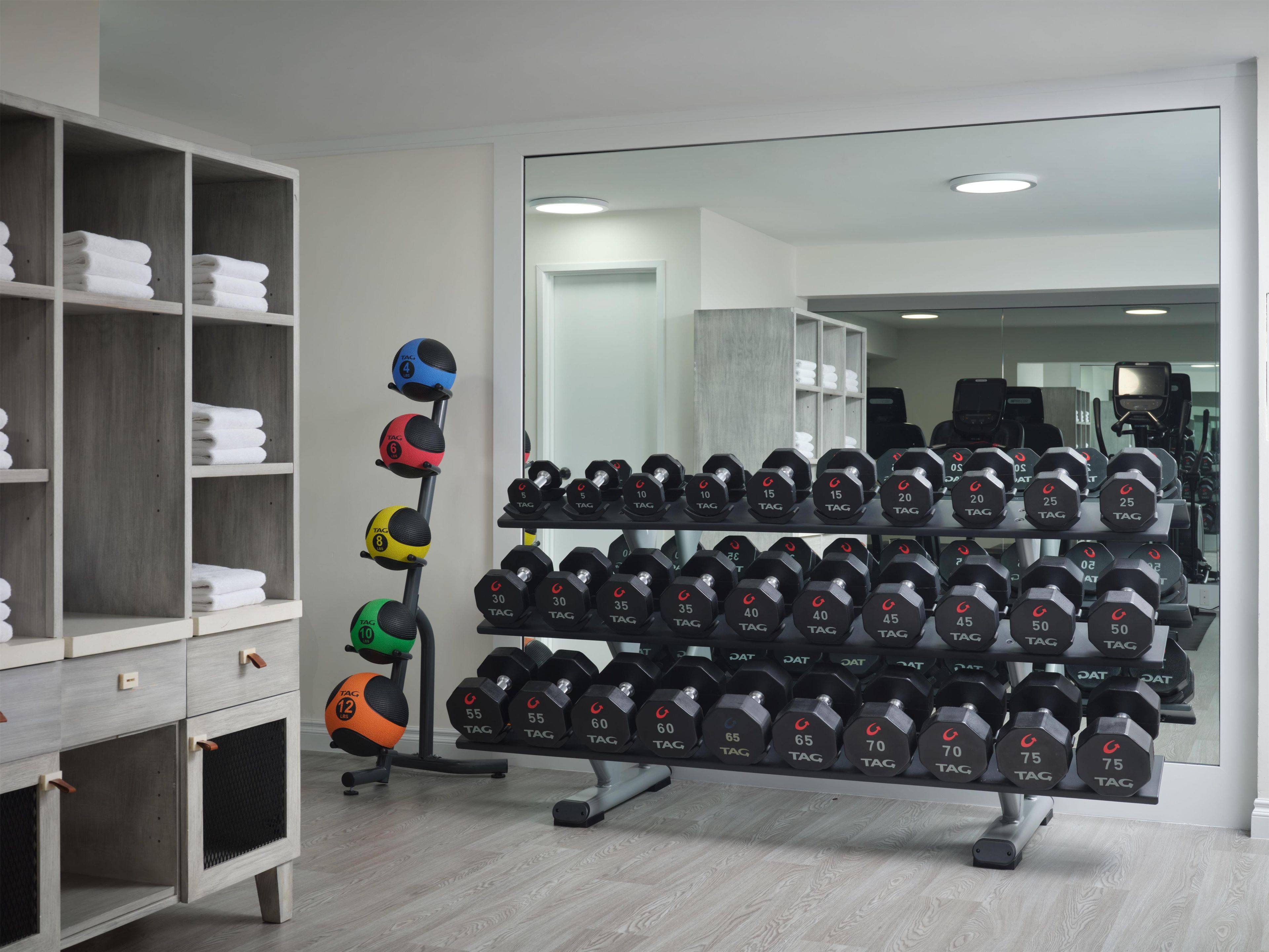 Un gimnasio con equipo de levantamiento de pesas, incluyendo mancuernas y balones medicinales, y un espejo que refleja el espacio.