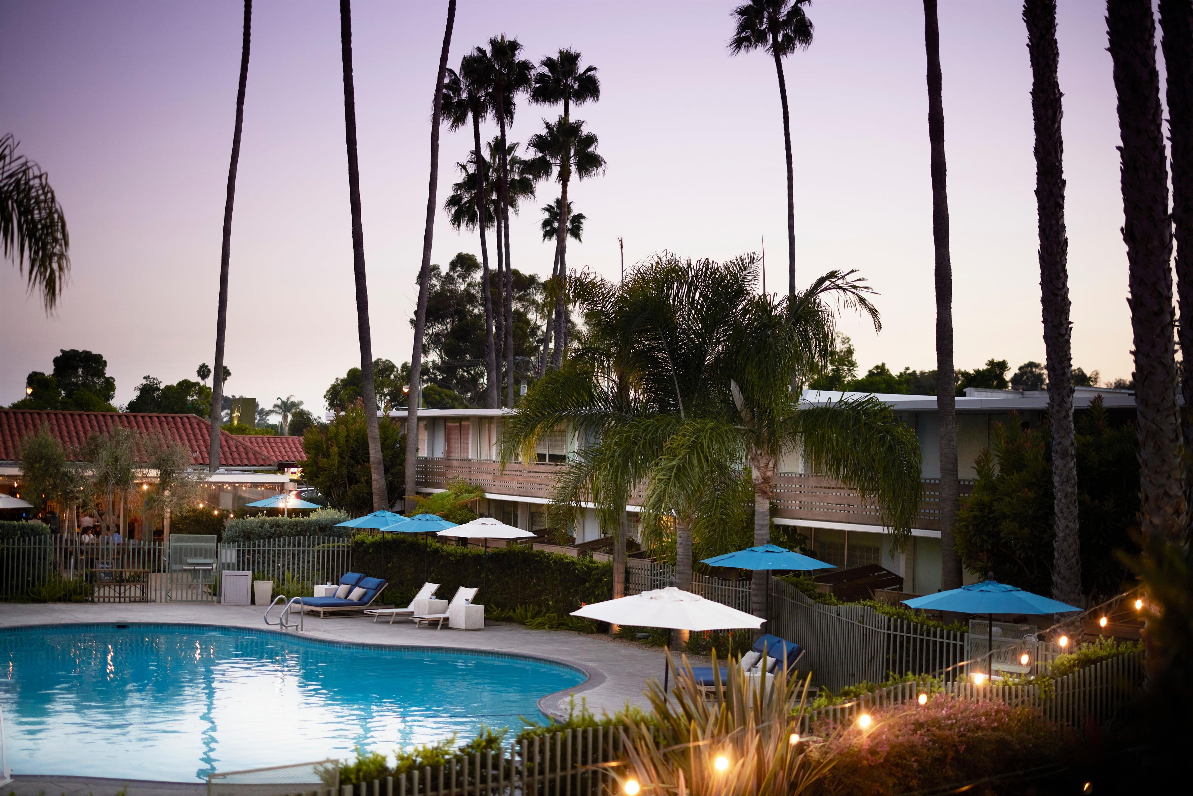 A swimming pool with lounge chairs and umbrellas, surrounded by palm trees and a building with a red roof.