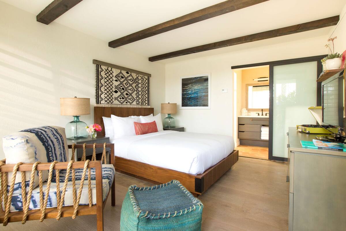 Une chambre avec un lit en bois, une chaise à dossier en corde, un petit pouf, une table de chevet avec une lampe et une commode avec un miroir.