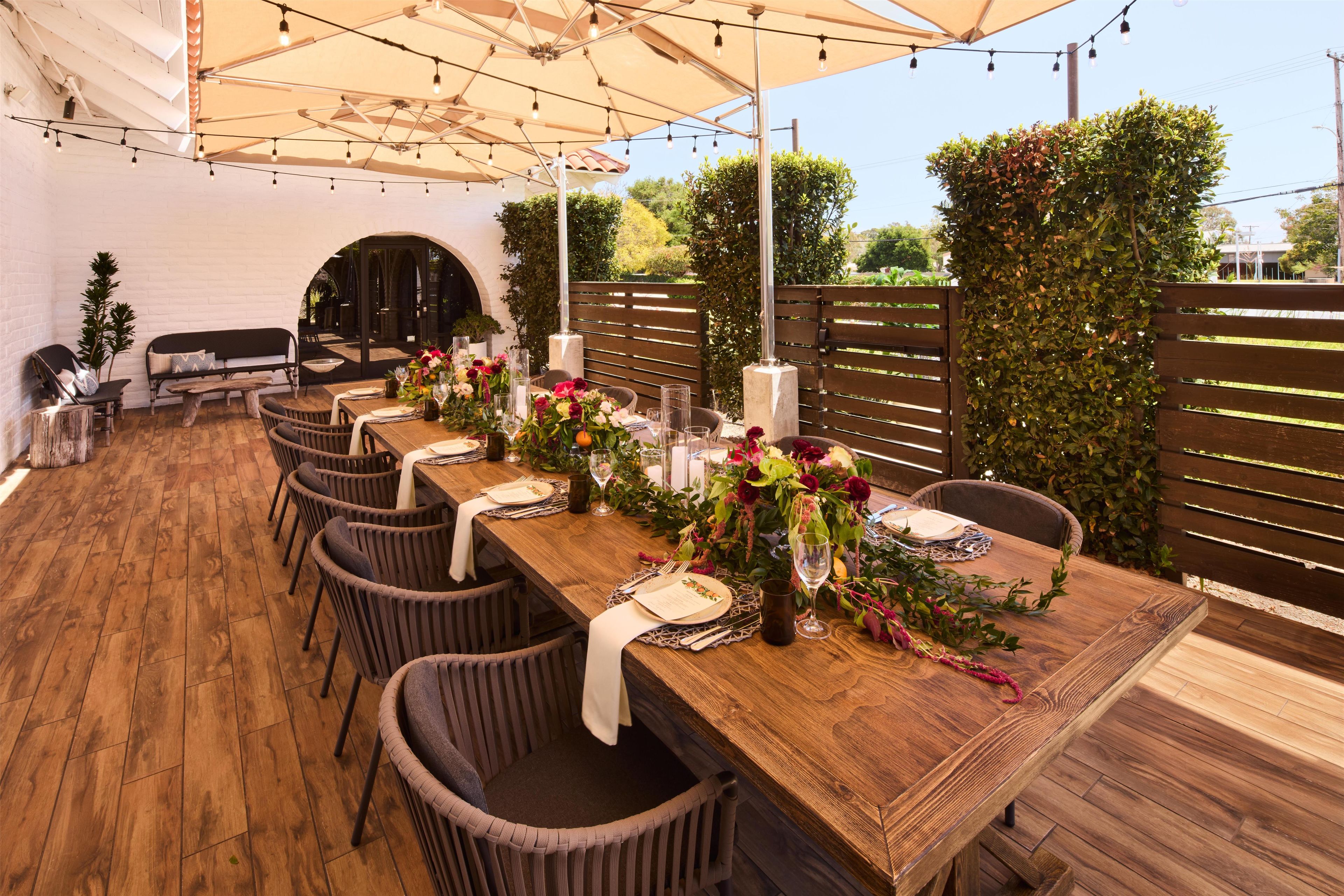 An outdoor dining area with a long wooden table set for a meal, surrounded by wicker chairs, under a canopy with string lights.