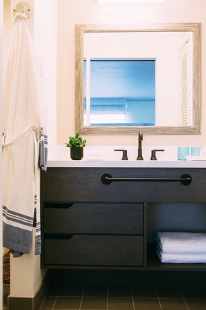 Un baño con un espejo grande, un lavabo con dos grifos, una pequeña planta en maceta y un perchero para toallas.