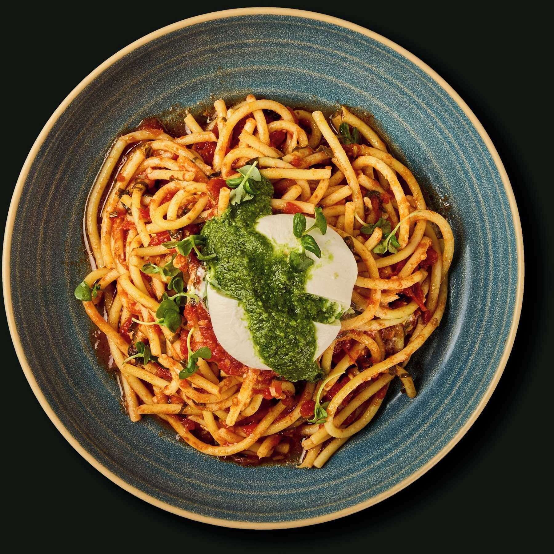 Un plato de espagueti con salsa de tomate, adornado con una cucharada de pesto y una loncha de mozzarella.