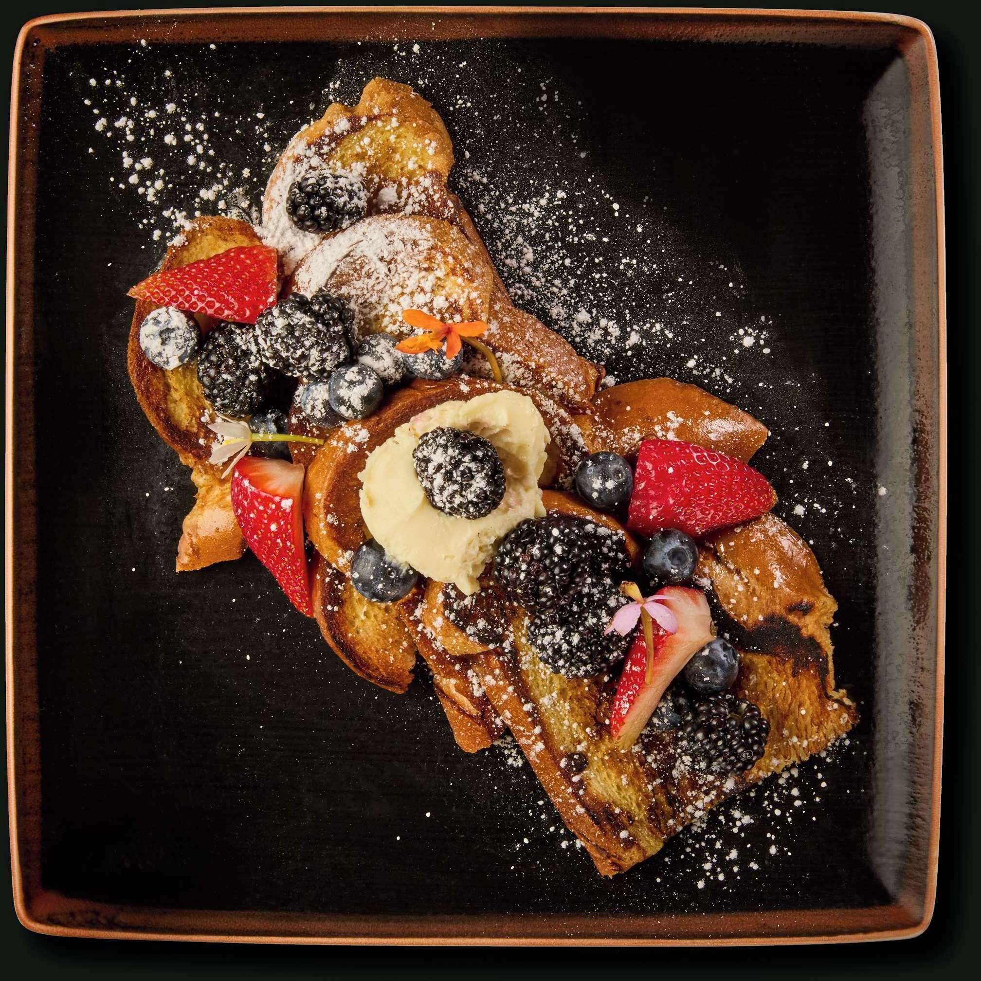 A square plate with a dark background, containing a stack of pancakes topped with strawberries, blueberries, blackberries, and a dollop of whipped cream.