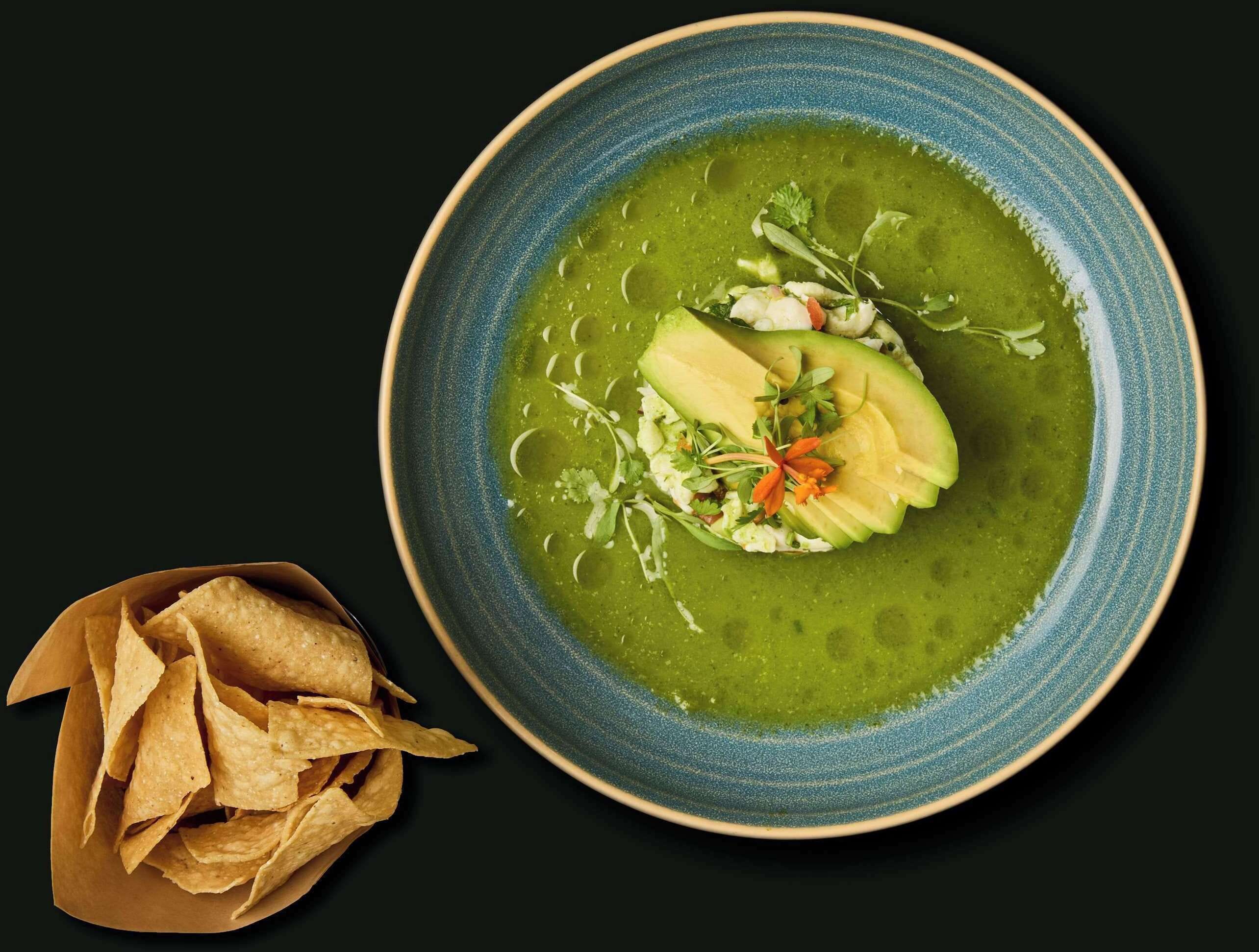 A bowl of green soup with avocado slices and garnishes, accompanied by tortilla chips.