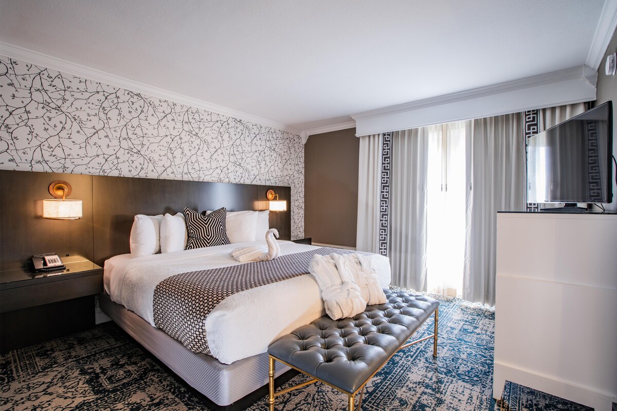 A hotel room with a large bed, a bedside table with a phone, a wall-mounted lamp, a TV, and a window with curtains.