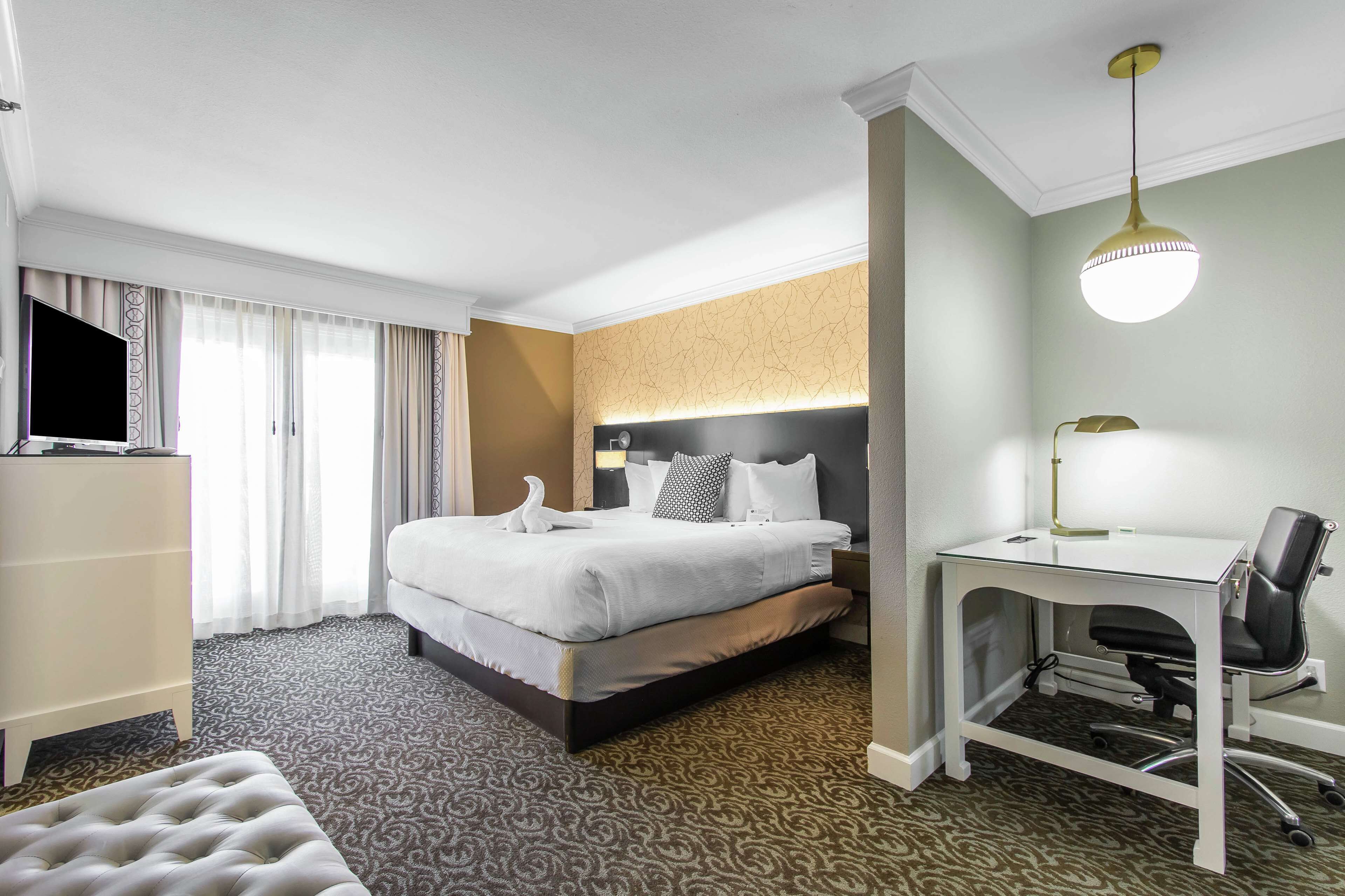 Quarto de hotel com cama, escrivaninha, cadeira, televisão e uma janela grande com cortinas.