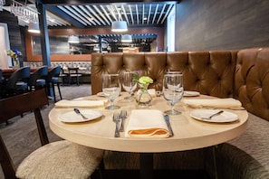A round dining table with a brown leather booth in the center, set with plates, cutlery, and wine glasses. - The Burgundy Hotel, Tapestry Collection by Hilton (Little Rock)