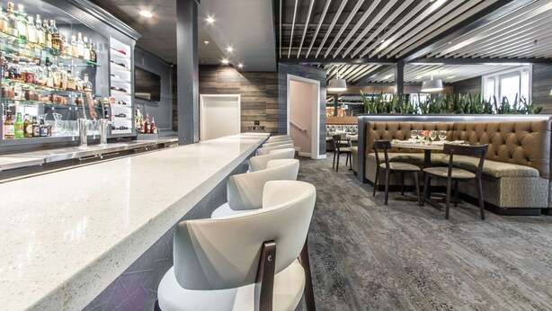 A modern bar area with a long counter, multiple bar stools, and a seating area with a cushioned bench and a table. - The Burgundy Hotel, Tapestry Collection by Hilton (Little Rock)