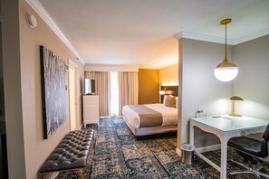 A hotel room with a bed, a desk with a chair, a bench, a trash can, and a wall-mounted TV. - The Burgundy Hotel, Tapestry Collection by Hilton (Little Rock)