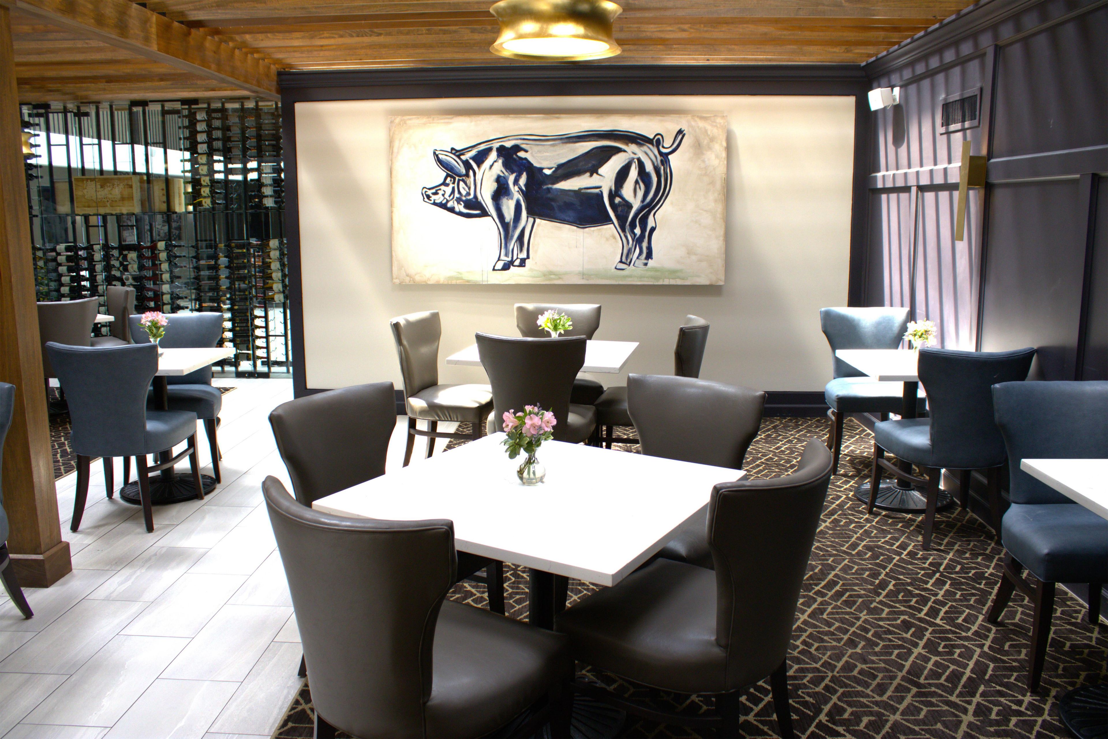 Interior de un restaurante con zona de comedor, mesas y sillas, una obra de arte en la pared con dos animales y una alfombra estampada.