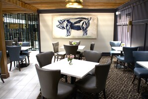 A restaurant interior with a dining area featuring tables and chairs, a wall-mounted artwork of two animals, and a patterned carpet. - The Burgundy Hotel, Tapestry Collection by Hilton (Little Rock)