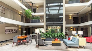 A spacious lobby with a high ceiling, a dining area with a table and chairs, a sofa, and a coffee table. There are plants and a large window in the center. - The Burgundy Hotel, Tapestry Collection by Hilton (Little Rock)